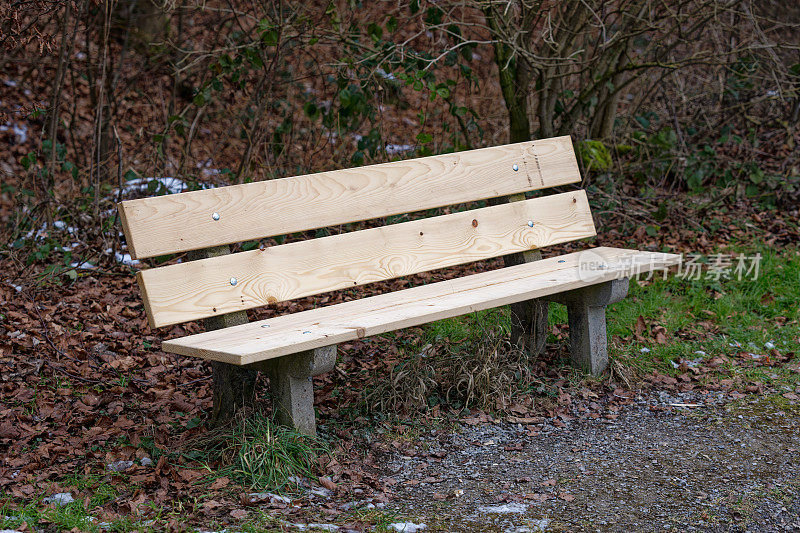 Empty wooden bench at border of gravel rural road at border of forest at City of Zürich district Schwamendingen on a gray and cloudy winter day.